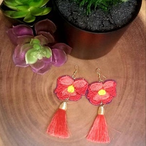 May include: A pair of red flower earrings with gold tassels. The earrings are on a wooden surface with green plants in the background.