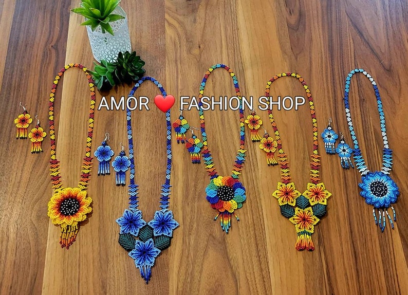 May include: Five sets of handcrafted beaded necklaces and matching earrings featuring vibrant floral designs in yellow, blue, and multi-colored patterns. The text AMOR ❤️ FASHION SHOP is displayed above the jewelry, which includes intricate beadwork.