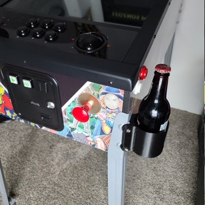 May include: A black pinball machine with a silver base and a black drink holder attached to the side. A bottle of beer is in the holder. The pinball machine has a red button and a black trackball.