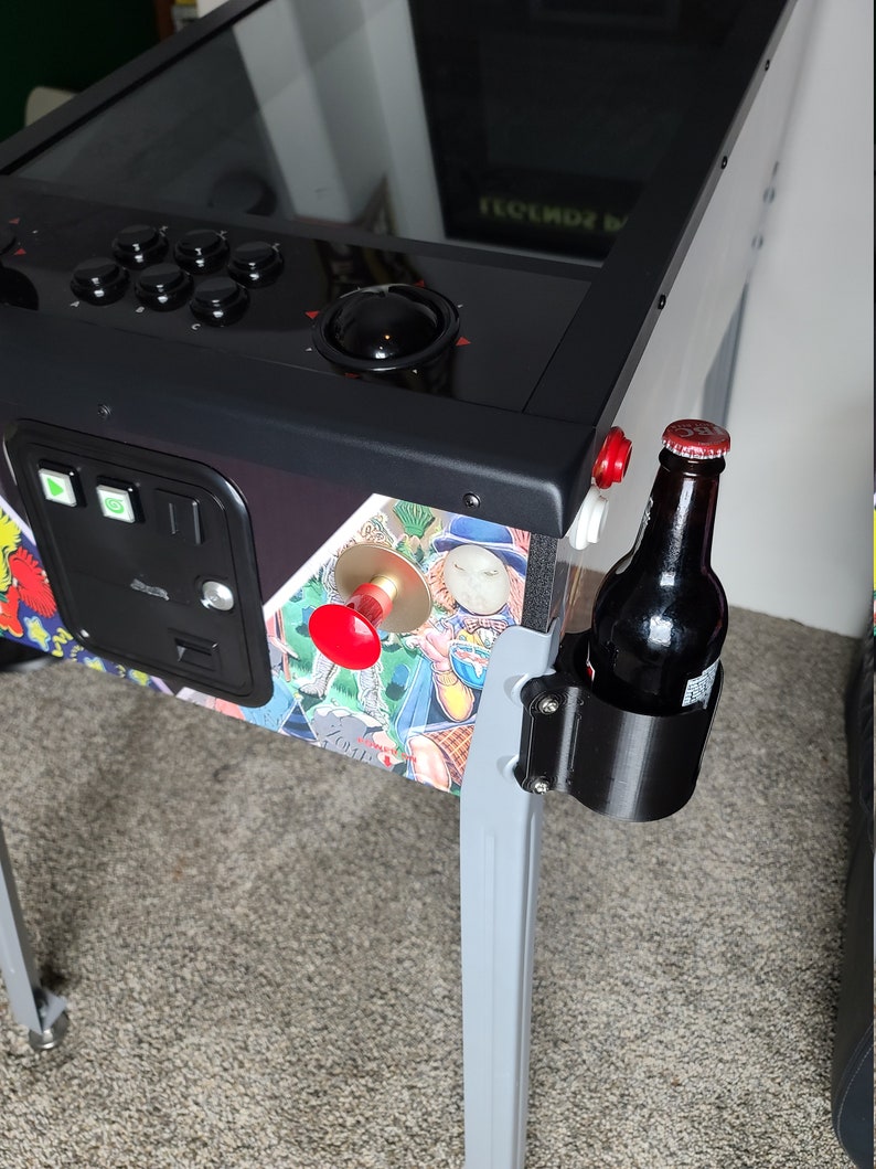 May include: A black pinball machine with a silver base and a black drink holder attached to the side. A bottle of beer is in the holder. The pinball machine has a red button and a black trackball.