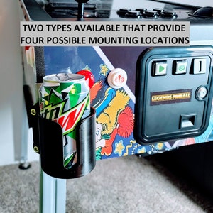 May include: A black plastic drink holder attached to a pinball machine. The holder is designed to hold a can of soda or a bottle of water. The text "TWO TYPES AVAILABLE THAT PROVIDE FOUR POSSIBLE MOUNTING LOCATIONS" is visible on the pinball machine.