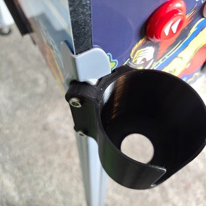 May include: A black plastic cup holder mounted on the side of a pinball machine. The cup holder is attached with a screw and has a round opening for a cup.