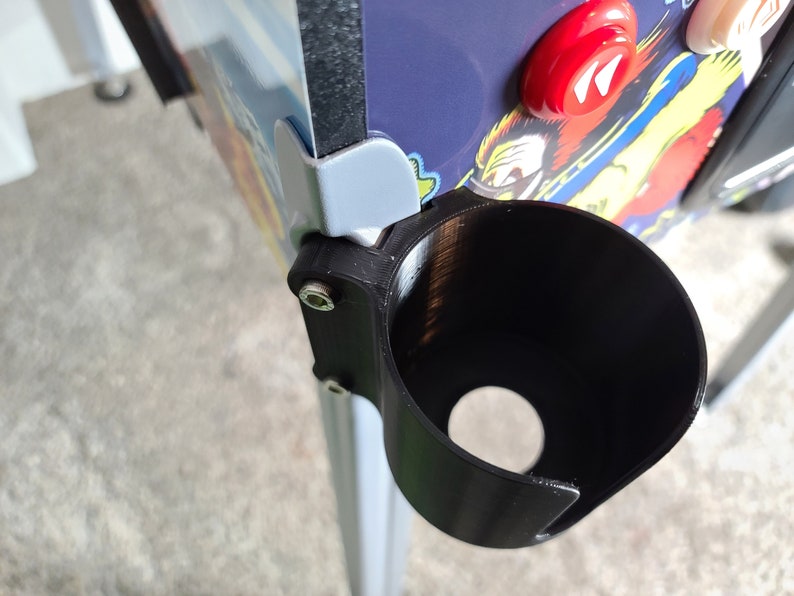 May include: A black plastic cup holder mounted on the side of a pinball machine. The cup holder is attached with a screw and has a round opening for a cup.