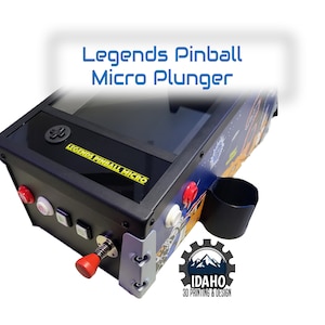 May include: A black and silver Legends Pinball Micro Plunger pinball machine with a red plunger, a cup holder, and a clear playing field. The machine has a yellow and black sticker that says "Legends Pinball Micro".