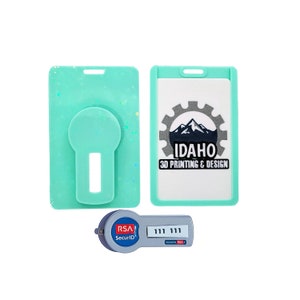 May include: Two turquoise blue plastic ID card holders, one with a clear window and the other with a round cutout for a photo. The holder with the window has a white background with a black and gray logo that says "IDAHO 3D PRINTING & DESIGN". A gray RSA SecurID token with a black display showing "111 111" is in the foreground.