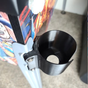 May include: A black plastic cup holder attached to a silver metal pole. The cup holder is designed to hold a beverage can or bottle.
