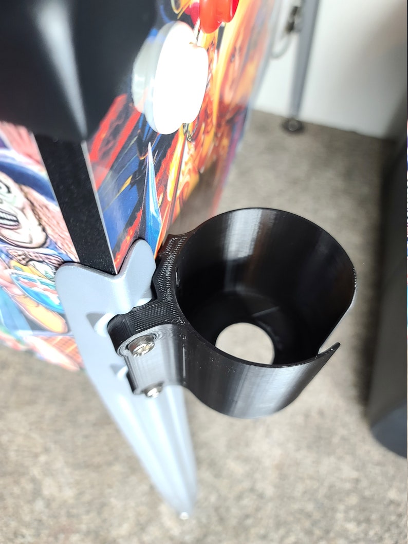 May include: A black plastic cup holder attached to a silver metal pole. The cup holder is designed to hold a beverage can or bottle.