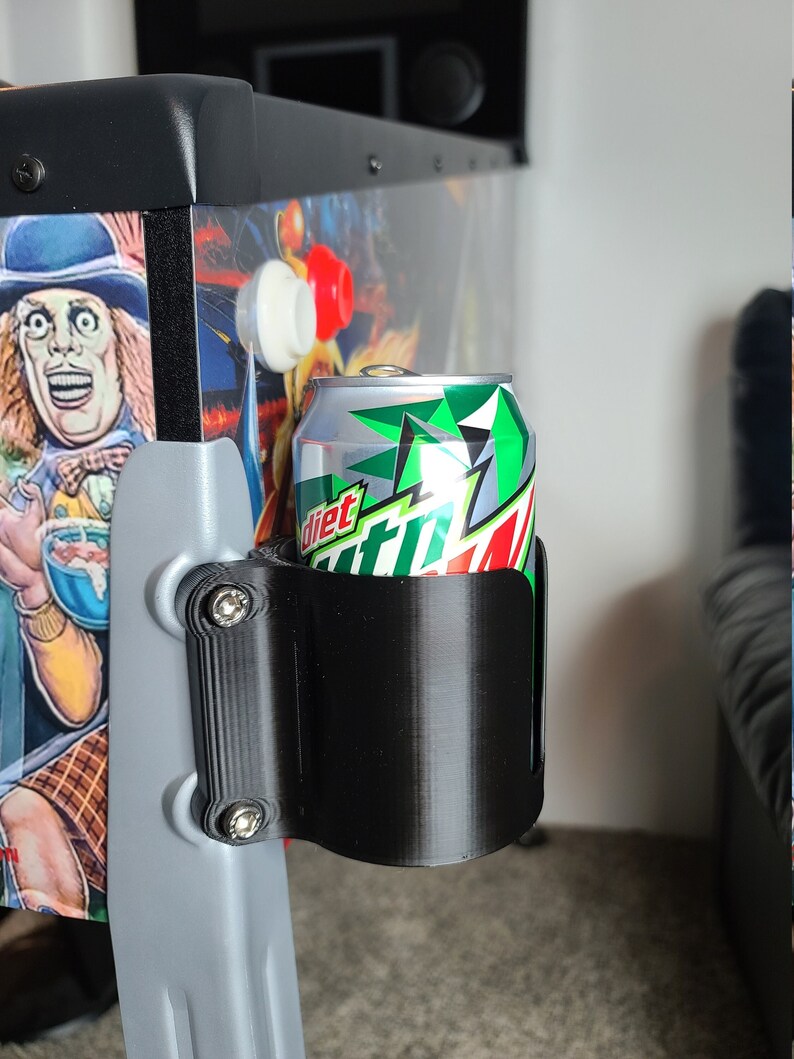 May include: A black 3D printed can holder attached to a pinball machine. A silver can with green and white labelling is in the holder. The can reads "Diet Mountain Dew."