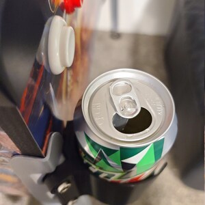 May include: A silver can with a green and white design is held in a black drink holder. The can has a pull tab and is partially visible.