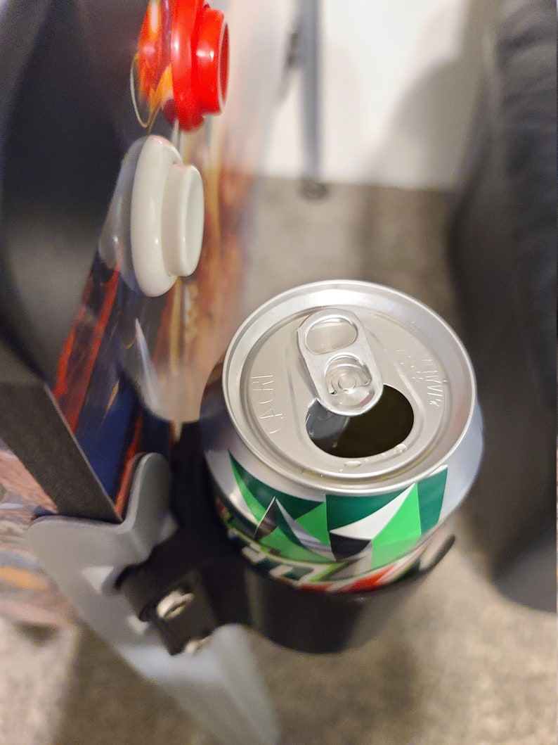 May include: A silver can with a green and white design is held in a black drink holder. The can has a pull tab and is partially visible.