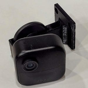 Puede incluir: Una cámara de seguridad exterior Blink negra montada en un soporte de pared de plástico negro. La cámara tiene una lente redonda y la palabra "blink" está impresa en la parte inferior.