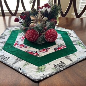 May include: A hexagonal table runner with a white background and a green border. The runner features a repeating pattern of red trucks and pine trees. The center of the runner is decorated with a green and white floral arrangement.