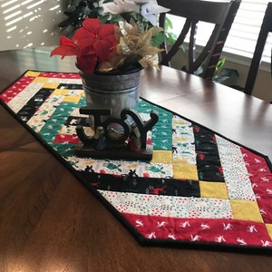 May include: A colorful quilted table runner with a black border, featuring a patchwork design of red, yellow, black, and white patterned fabric. A galvanized metal bucket with red, white, and gold flowers sits on the runner, with a wooden "JOY" sign.
