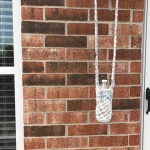 May include: A white and blue crocheted water bottle holder with a long strap, hanging against a red brick wall. The holder contains a clear plastic water bottle. The strap is made of the same crocheted material.