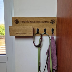 May include: Wooden wall-mounted dog leash and collar holder with three hooks. The holder has a sign that reads "Time to walk the human" with paw prints on either side. A small shelf is below the sign with the words "Dora Cocker" engraved on it.