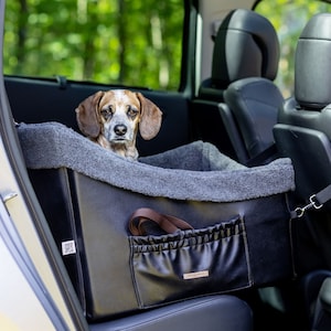 Puede incluir: Un asiento de coche para mascotas negro con un forro de vellón gris. Un perro marrón está sentado en el asiento, mirando a la cámara. El asiento tiene un bolsillo con cremallera y un enganche para la correa.
