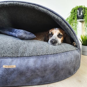 May include: A cozy dog bed with a built-in hood in shades of gray. The bed features a plush interior and a faux leather exterior. A small dog is nestled inside, with a blue plush toy resting on the bed. The brand name "Dora Cocker" is visible on a label.