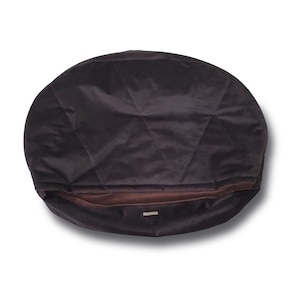 Puede incluir: Un puf redondo de color marrón oscuro con la parte superior acolchada y una cremallera marrón. El puf está hecho de un tejido suave y mullido.