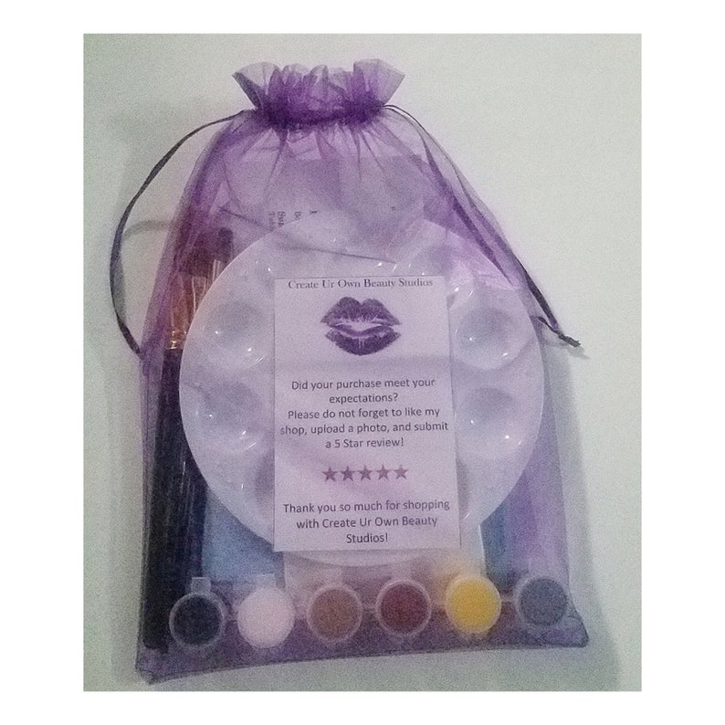 Puede incluir: Una bolsa de organza morada llena de suministros de pintura para un proyecto de arte de labios. La bolsa incluye una paleta blanca con seis pozos, cinco peque&ntilde;os botes de pintura, dos pinceles y una tarjeta que dice "Create Ur Own Beauty Studios" con un dise&ntilde;o de labios y una solicitud para dejar una rese&ntilde;a.