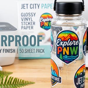 May include: A clear water bottle covered in colorful stickers with the text "Explore PNW." The bottle is next to a box of glossy vinyl sticker paper and a printer. The text "Waterproof Vinyl Glossy Finish 50 Sheet Pack" is also visible.