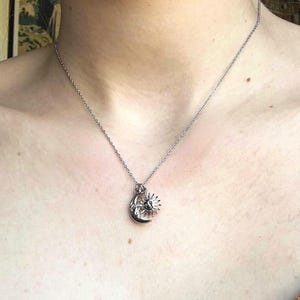 May include: Silver-toned necklace featuring a pendant with a crescent moon and a sunburst design. The pendant is suspended from a delicate chain. The necklace is worn against a brown top.