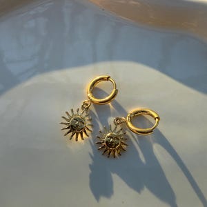 以下が含まれることがあります： 金色のフープピアスのペア。それぞれのフープから小さな太陽のチャームがぶら下がっています。太陽のチャームには顔が描かれています。