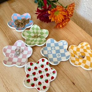 Puede incluir: Una colección de cuencos de cerámica con forma de flor en varios diseños y colores, incluyendo diseños a cuadros y de fresas. Algunos cuencos contienen joyas, como collares y anillos. Los cuencos están colocados sobre una superficie de madera.