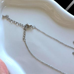 May include: A silver-colored necklace with a delicate chain and a small pendant. The necklace is displayed on a white surface, showcasing its intricate design and reflective qualities. The chain has an adjustable length.