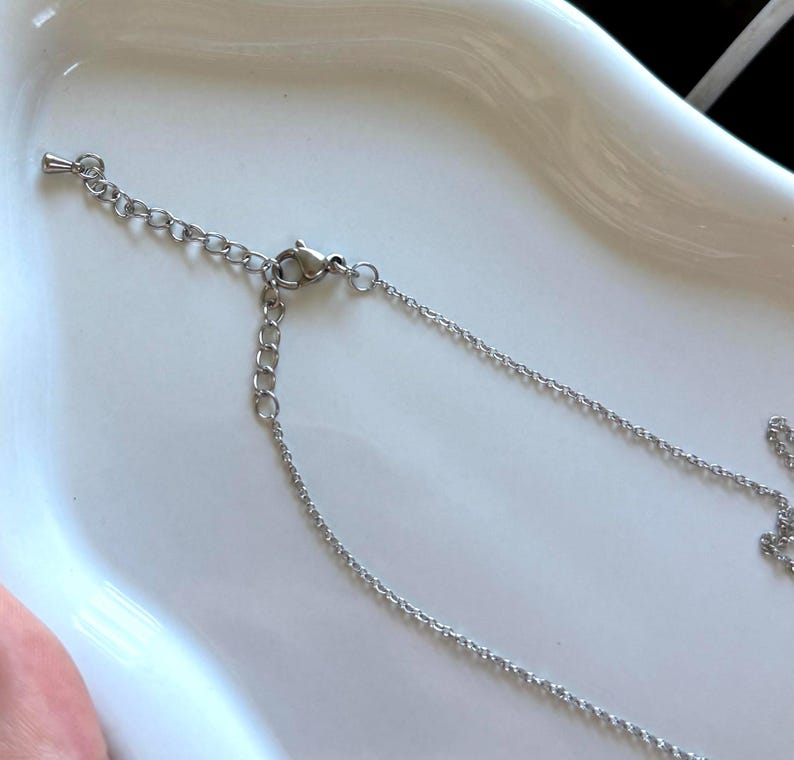 May include: A silver-colored necklace with a delicate chain and a small pendant. The necklace is displayed on a white surface, showcasing its intricate design and reflective qualities. The chain has an adjustable length.