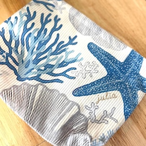 Op de afbeelding: Gevouwen stof met een maritiem thema. Het ontwerp bevat blauwe koraal-, zeester- en schelpenillustraties op een crèmekleurige achtergrond. De naam "julia" is in gouden script op de stof gedrukt.