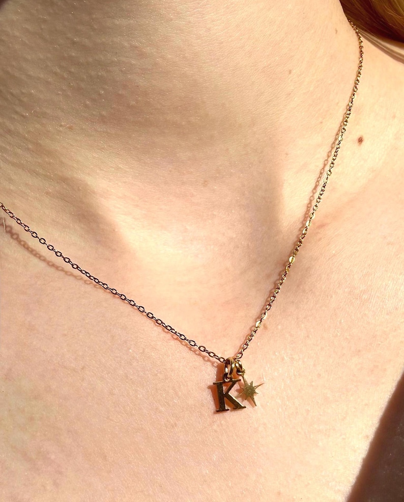 May include: A gold-colored necklace with a delicate chain. The pendant features the letter "K" and a small star charm. The necklace is worn around the neck.