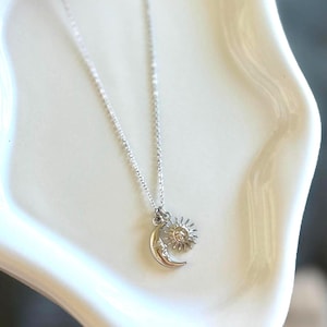 May include: A silver-colored necklace featuring a crescent moon and sun pendant. The moon has a face and the sun has rays. The necklace is displayed on a white, curved surface.