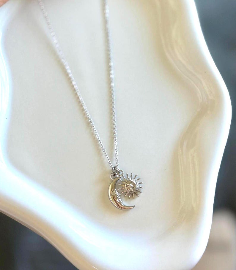 May include: A silver-colored necklace featuring a crescent moon and sun pendant. The moon has a face and the sun has rays. The necklace is displayed on a white, curved surface.