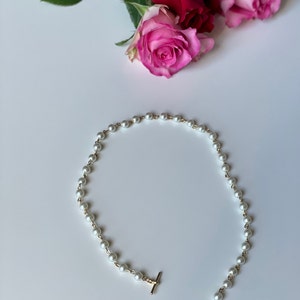 May include: A delicate pearl necklace with a gold clasp and a small gold ring at the end. The necklace is on a white surface.