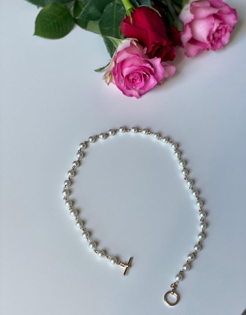 May include: A delicate pearl necklace with a gold clasp and a small gold ring at the end. The necklace is on a white surface.