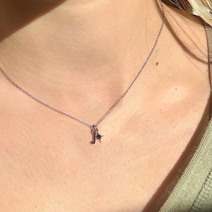 May include: A silver necklace with a delicate chain. The pendant features the letter "J" and a small star charm. The necklace is worn around the neck, with the chain resting on the skin.