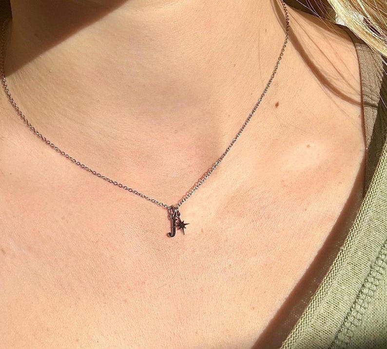 May include: A silver necklace with a delicate chain. The pendant features the letter "J" and a small star charm. The necklace is worn around the neck, with the chain resting on the skin.
