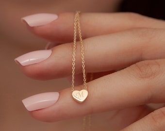 May include: A delicate gold necklace featuring a heart-shaped pendant. The pendant is engraved with the letter "M". The necklace is held by a person with manicured nails.