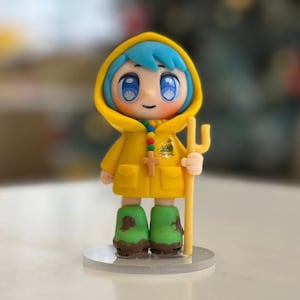 Könnte beinhalten: Figur einer Figur mit blauen Haaren, die einen gelben Kapuzenmantel und grüne Stiefel trägt. Die Figur hält einen Stab und steht auf einer durchsichtigen Basis. Der Mantel hat einen Kreuzanhänger.