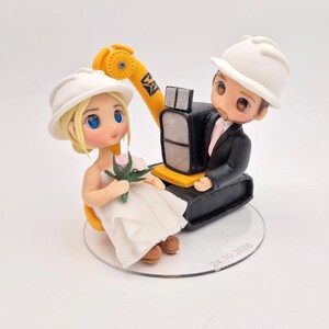 May include: A cake topper featuring two small figurines wearing white hard hats. One figure wears a white dress and holds a small flower, while the other wears a black suit and sits on a yellow and black toy excavator. The date 24.10.2028 is printed on the clear base.