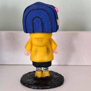 May include: A figurine of a character with blue hair, a yellow jacket, and black trousers. The figurine has a pink hair accessory and is standing on a textured, dark grey base. The figure's boots are yellow with white stripes.
