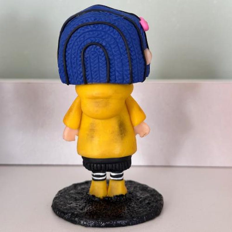 May include: A figurine of a character with blue hair, a yellow jacket, and black trousers. The figurine has a pink hair accessory and is standing on a textured, dark grey base. The figure's boots are yellow with white stripes.