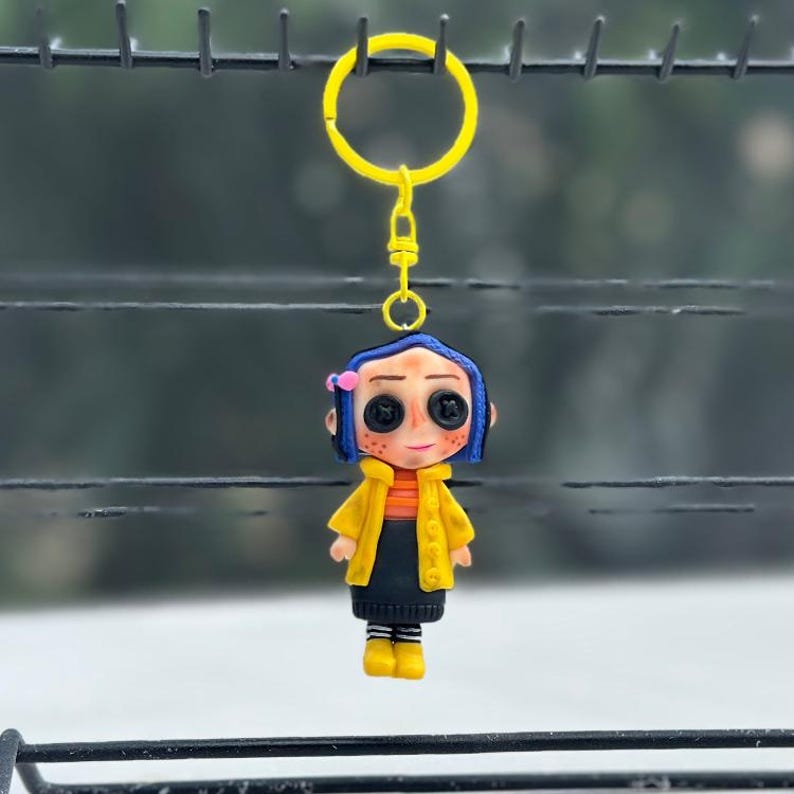 May include: A handcrafted keyring featuring a doll-like character with button eyes, blue hair, and a yellow jacket. The figure is attached to a yellow metal keyring. The doll is wearing a black skirt and yellow boots. The background is blurred.