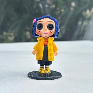 A small, yellow raincoat with black buttons and a blue wig with a pink bow. The figure has large, black button eyes and a black skirt. The figure is standing on a black base.