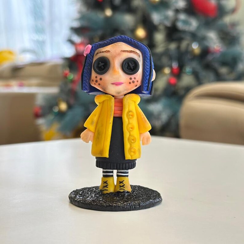 May include: A figurine of a character with blue hair, button eyes, and freckles, wearing a yellow coat over an orange striped shirt and black skirt. The figurine stands on a textured black base, with yellow boots. The background is blurred.