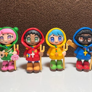 Könnte beinhalten: Vier handgefertigte Figuren von Kindern, die verschiedene farbige Kleidung tragen und eine Heugabel halten. Die Kinder tragen grüne, rote, gelbe und blaue Kleidung. Jede Figur hat ein Kreuz auf der Brust.