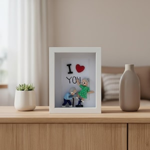 Puede incluir: Una obra de arte enmarcada con las palabras "I ❤️ YOU" y una representación de una pareja de ancianos. La mujer viste un vestido verde, el hombre una camisa azul. Una pequeña maceta con suculentas y un jarrón están sobre una superficie de madera.