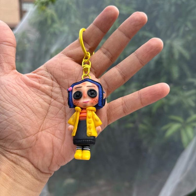 May include: A handmade keychain featuring a doll-like figure with button eyes, blue hair, and a yellow coat. The figure has an orange and black outfit and yellow boots. The keychain is attached to a yellow metal ring.