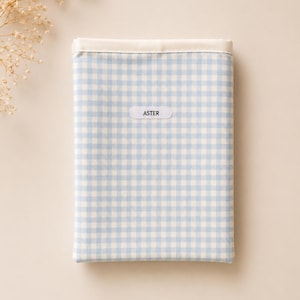 Può includere: Un tessuto rettangolare con un motivo a quadretti vichy blu chiaro e bianco, con un bordo bianco. La parola "ASTER" è stampata su una piccola etichetta bianca, centrata sul tessuto. Il tessuto è posto su uno sfondo neutro, con fiori secchi nell'angolo in alto a sinistra.