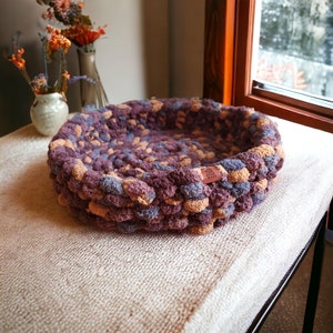 May include: A round, purple and tan crocheted pet bed with a brown leather tag that says "Hand Made".
